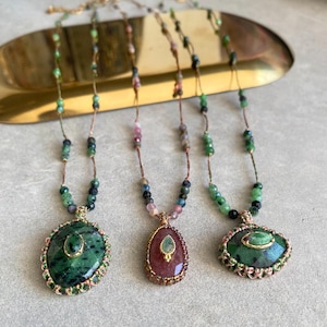 May include: Three gemstone necklaces with different colored pendants and beaded chains. The pendants are oval and teardrop shaped, with green, pink, and multi-colored stones. The necklaces are displayed on a gold-colored tray.