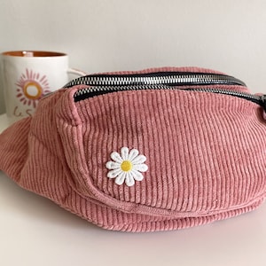 Op de afbeelding: Een roze corduroy heuptas met een zwarte rits en een witte gestikte madeliefje op de voorkant.