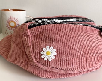 May include: A pink corduroy fanny pack with a black zipper and a white daisy embroidered on the front.