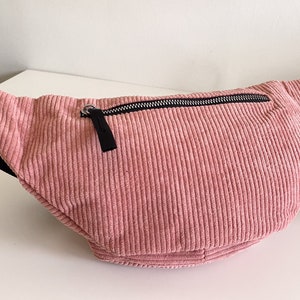 Op de afbeelding: Een roze corduroy heuptas met een zwarte rits en een zilveren rits-trekker.