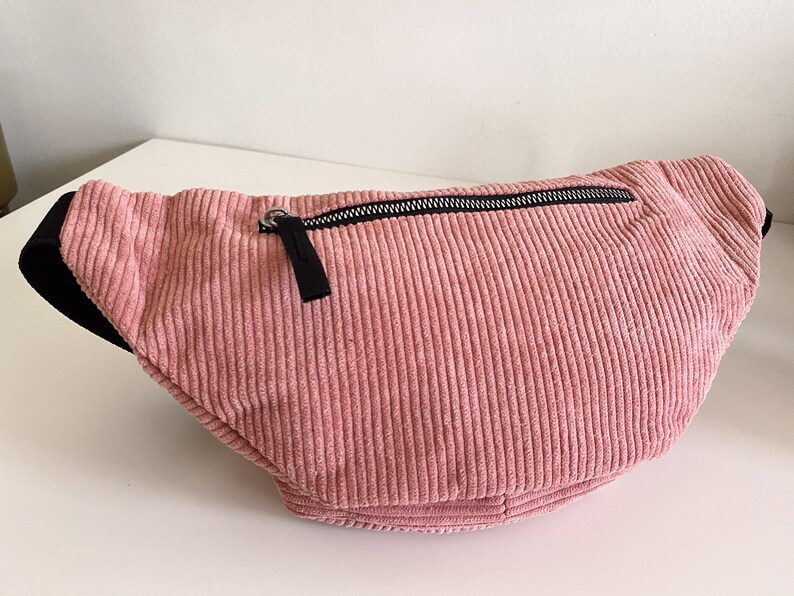 Op de afbeelding: Een roze corduroy heuptas met een zwarte rits en een zilveren rits-trekker.