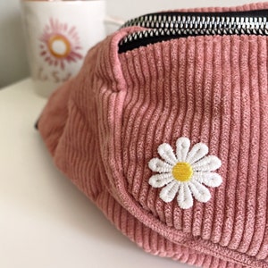 Op de afbeelding: Een roze corduroy heuptas met een zwarte rits en een witte gestikte madeliefje op de voorkant.
