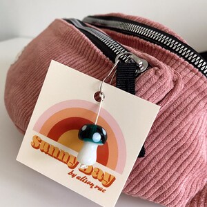 Op de afbeelding: Een roze corduroy heuptas met een zwarte rits en een label met de tekst "Sunny Day by alison rae". Een witte en groene paddenstoel-hanger is aan de rits bevestigd.
