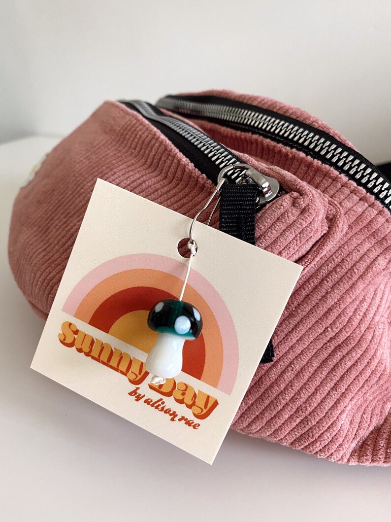 Op de afbeelding: Een roze corduroy heuptas met een zwarte rits en een label met de tekst "Sunny Day by alison rae". Een witte en groene paddenstoel-hanger is aan de rits bevestigd.
