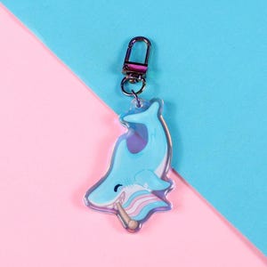 May include: A light blue shark keychain with a silver clasp. The shark is holding a flag with pink, white, and blue stripes. The background is split diagonally with pink and blue.