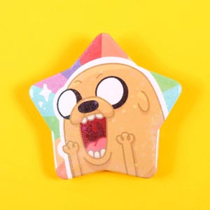 May include: A star-shaped badge featuring a cartoon dog with a surprised expression. The badge has a rainbow-coloured border and a yellow background. The dog has large black eyes and a wide-open mouth.