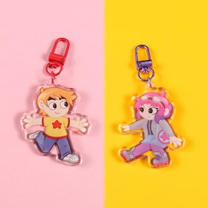 May include: Two acrylic keychains featuring cartoon characters. One keychain depicts a boy in a yellow shirt with a red star, blue pants, and white shoes. The other keychain shows a girl with pink hair, goggles, and a gray jacket, on a pink and yellow background.