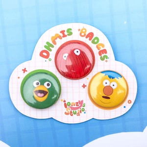 Puede incluir: Un conjunto de tres chapas circulares con caras de personajes sobre una tarjeta de soporte en forma de nube. La tarjeta muestra el texto DHMIS BADGES e ionzy's STUDIO. Las chapas incluyen un personaje rojo, uno verde tipo pájaro y uno amarillo.