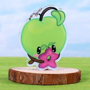 Könnte beinhalten: Ein klarer Acryl-Schlüsselanhänger mit einem Cartoon-Apfel-Charakter mit grünem Körper, schwarzen Augen und einer rosa sternförmigen Blume. Der Schlüsselanhänger ist an einer silbernen Metallkette und einem Ring befestigt. Der Apfel wird auf einer kleinen Holzscheibe präsentiert.