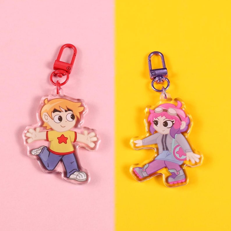 May include: Two acrylic keychains featuring cartoon characters. One keychain depicts a boy in a yellow shirt with a red star, blue pants, and white shoes. The other keychain shows a girl with pink hair, goggles, and a gray jacket, on a pink and yellow background.