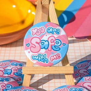 Puede incluir: Un botón redondo con el texto "You are Safe with me" en rosa y azul, exhibido en un pequeño caballete de madera. El botón tiene un fondo a rayas rosa y azul con detalles de corazón. Se ven otros botones.