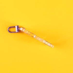 May include: A clear acrylic keychain with a silver-colored clasp, lying diagonally on a bright yellow background. The keychain has a rectangular shape with etched details. The clasp is a simple, sturdy design.