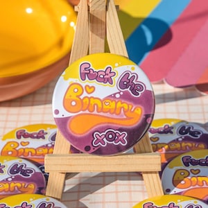 May include: A circular badge with the words "Fuck the Binary" in white and orange, set against a yellow and purple backdrop. The badge is displayed on a small wooden easel, with other similar badges visible.