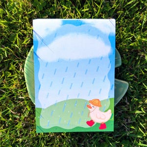 Puede incluir: Bloc de notas rectangular con una ilustración de un pato con sombrero y botas de lluvia, sobre un fondo de cielo lluvioso y paisaje verde. El bloc de notas presenta un diseño de nubes azules y blancas.