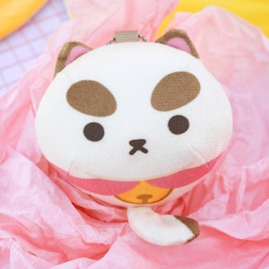 May include: A soft cat-shaped purse, white with brown markings, a pink collar, and a gold bell. The cat has brown ears and a small tail. It rests on a pink, textured paper background.