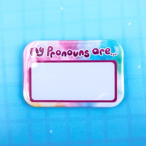 May include: A rectangular name badge with rounded corners featuring a watercolor-style background in shades of pink, blue, and yellow. The text "my pronouns are..." is printed in dark purple, above a large, blank white rectangular space.