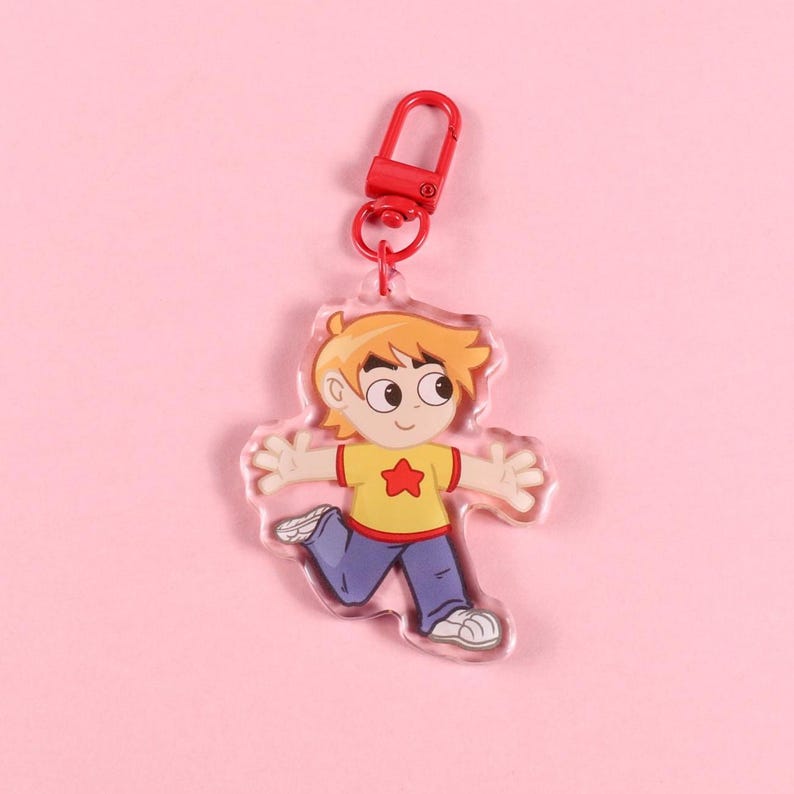 May include: A clear acrylic keychain featuring a cartoon boy with orange hair, a yellow shirt with a red star, blue pants, and white shoes. The keychain has a red clasp and a red ring. The background is a solid pink.