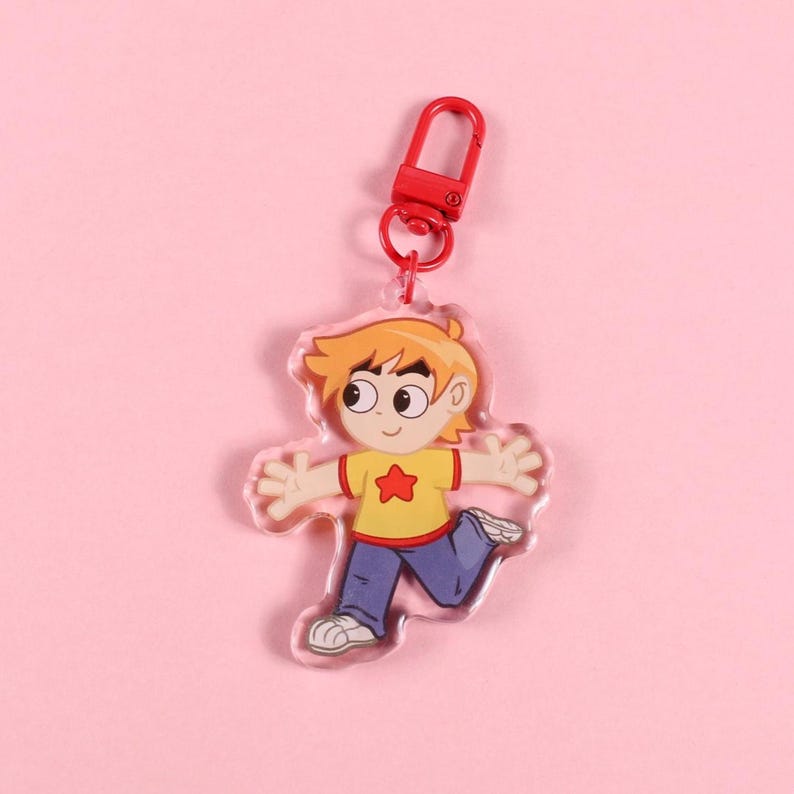 May include: A clear acrylic keychain featuring a cartoon boy in a yellow shirt with a red star, blue pants, and white shoes. The keychain has a red clasp and a red ring. The boy is in a running pose with his arms outstretched. The background is pink.