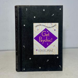 Può includere: Un libro nero con un designo a forma di diamante viola e giallo sulla copertina. Il titolo del libro è "Get Psychic!" e l'autore è Stacey Wolf. La copertina del libro è decorata con stelle bianche e punti interrogativi.