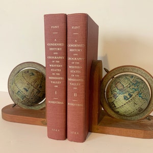 Peut inclure: Deux livres reliés en rouge intitulés "A Condensed History and Geography of the Western States or the Mississippi Valley" par Flint Rosenthal. Les livres sont des serre-livres avec un globe de chaque côté.