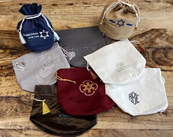 May include: An assortment of drawstring bags in various colors, including navy, gray, and burgundy. Each bag features embroidered designs such as a Star of David, leaves, and Hebrew text. Some bags have tassels.