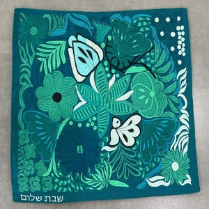 May include: Embroidered square textile in shades of teal, turquoise, and white, featuring a floral design. The design includes various flowers and leaves. The words "Shabbat Shalom" are embroidered in white at the bottom left corner.
