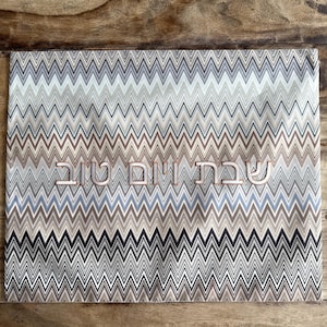 May include: A rectangular fabric cover featuring a repeating chevron pattern in shades of beige, brown, grey, white, and black. The center of the fabric displays the Hebrew text שבת ויום טוב embroidered in a light-colored thread with a thin outline.