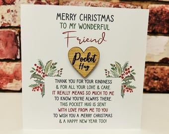 Könnte beinhalten: Eine weiße Weihnachtsgrußkarte mit einem hölzernen Herz-Token und der Aufschrift Pocket Hug. Die Karte enthält den Text Merry Christmas to my wonderful friend, gefolgt von einem Gedicht über Freundlichkeit und Liebe, verziert mit Stechpalmen.