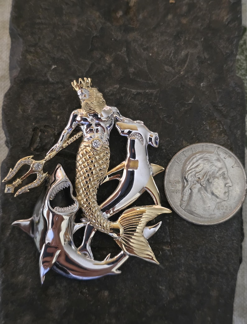 May include: A two-tone metal pendant featuring a mythical merman with a crown and trident, riding two sharks. The pendant has gold and silver tones with a diamond accent. A U.S. quarter is included for size reference.