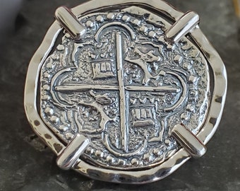 May include: Silver pendant in the shape of a Spanish eight reales coin with a cross and ornate design. The pendant is attached to a silver chain.