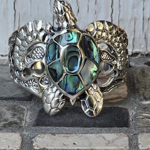 May include: A silver turtle bracelet with a green and blue abalone shell inlay. The turtle is detailed with textured scales and a raised head.