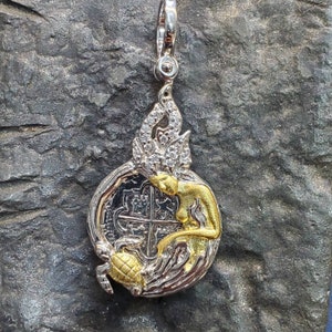 May include: Silver and gold pendant featuring a mermaid holding a silver coin with a cross design. The mermaid's hair is adorned with white stones and a turtle is at the bottom of the pendant.