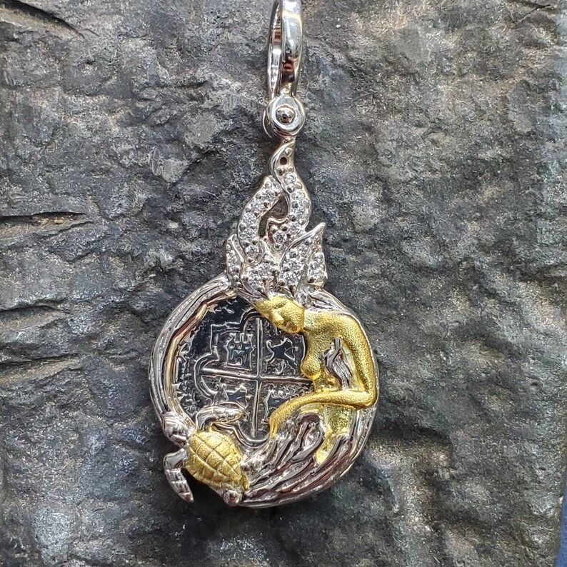 May include: Silver and gold pendant featuring a mermaid holding a silver coin with a cross design. The mermaid's hair is adorned with white stones and a turtle is at the bottom of the pendant.