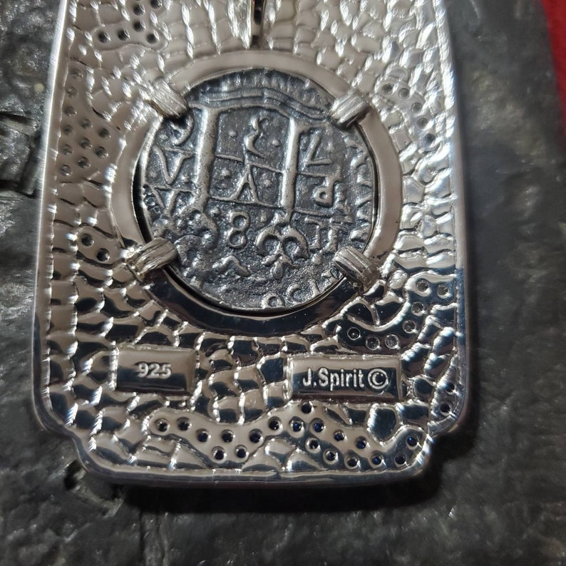 May include: A silver pendant featuring a replica of a Spanish silver coin with the inscription "8 Reales" and the year "1780". The pendant is marked "925" and "J. Spirit &copy;".