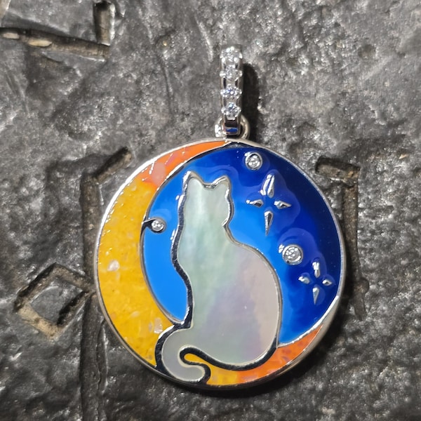 Pode incluir: Um pingente de prata com uma silhueta de gato branco em uma lua crescente azul e amarela. O pingente é decorado com pequenos strass transparentes.
