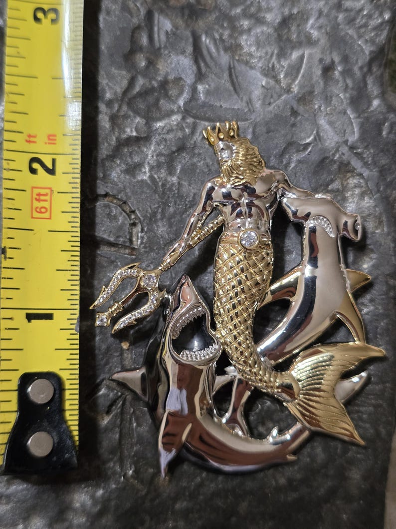 May include: A two-tone metal pendant featuring a gold-toned merman with a crown and a diamond-like gem, holding a trident. The merman is flanked by two silver-toned sharks. The pendant measures approximately 2 inches in height.