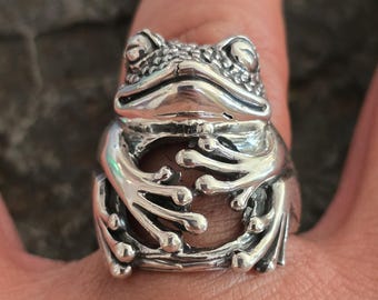 May include: A silver frog ring with detailed sculpting. The frog is depicted with its arms crossed in front of its body.