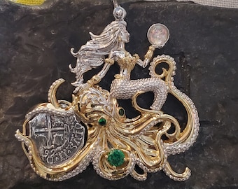 Könnte beinhalten: Ein silberner und goldener Anhänger mit einer Meerjungfrau, die auf einem Kraken reitet. Die Meerjungfrau hält eine perlmuttartige Kugel in ihrer rechten Hand. Der Kraken hat grüne Edelsteine an seinen Tentakeln. Der Anhänger befindet sich auf einem schwarzen Hintergrund.