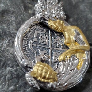 May include: A silver and gold pendant featuring a stylized mermaid with a gold tail, a turtle with a gold shell, and a silver coin with a coat of arms.