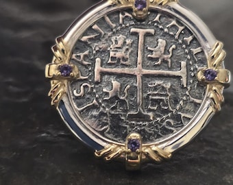 May include: Silver and gold pendant featuring a replica of an old Spanish coin with a cross and four lions. The pendant is accented with four purple gemstones.