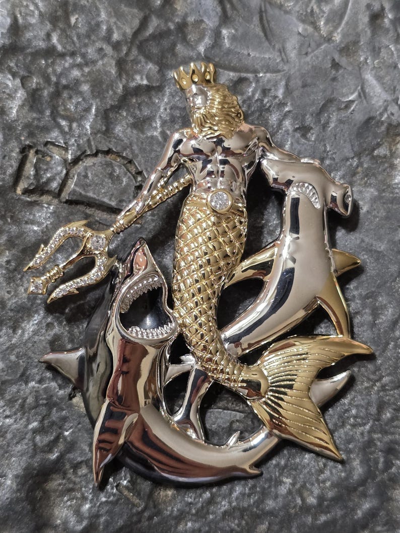 May include: A detailed pendant featuring a gold and silver merman with a crown, holding a jeweled trident. The merman is surrounded by two hammerhead sharks and a shark with an open mouth. The pendant has a diamond in the center.