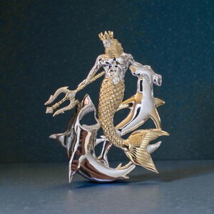 May include: A detailed, two-tone metal sculpture of a merman with a trident, crown, and a diamond accent. The merman is surrounded by two sharks. The sculpture is gold and silver in color.