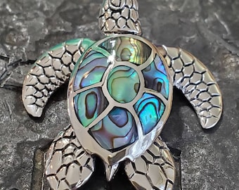 Può includere: Un ciondolo a forma di tartaruga marina argentata con intarsio di conchiglia di abalone iridescente. Il guscio presenta sfumature di blu, verde e viola. La testa, le pinne e i bordi del guscio della tartaruga hanno un design strutturato.
