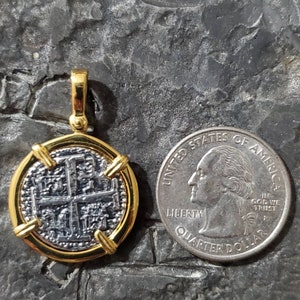 Pu&ograve; includere: Un ciondolo placcato oro con una replica in argento di una moneta spagnola da otto reali. La moneta ha un design a croce ed &egrave; circondata da una cornice dorata. Il ciondolo &egrave; mostrato accanto a una moneta da 25 centesimi di dollaro americano per il confronto delle dimensioni.