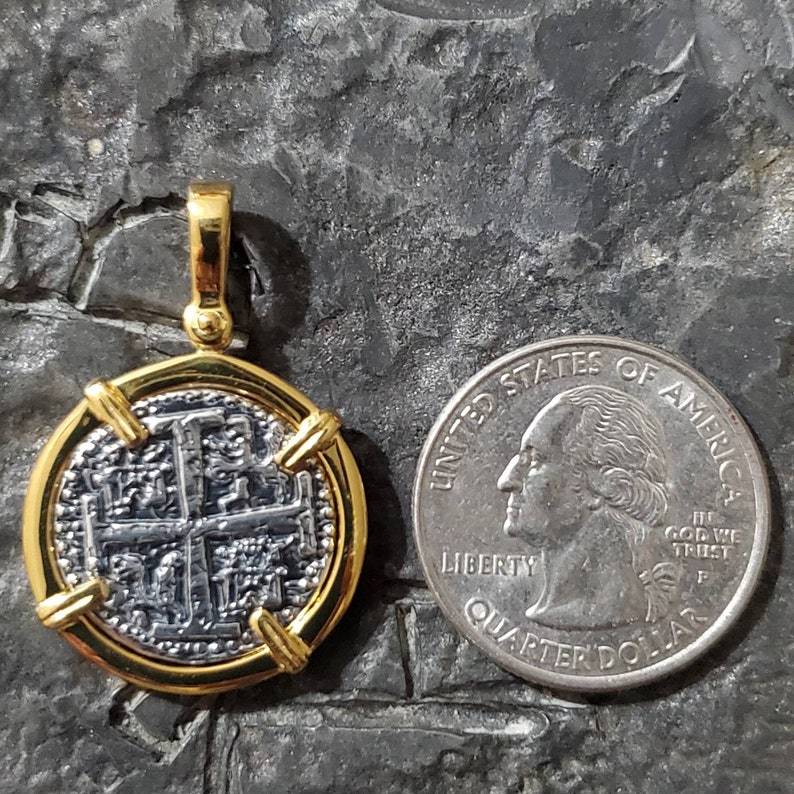 Pu&ograve; includere: Un ciondolo placcato oro con una replica in argento di una moneta spagnola da otto reali. La moneta ha un design a croce ed &egrave; circondata da una cornice dorata. Il ciondolo &egrave; mostrato accanto a una moneta da 25 centesimi di dollaro americano per il confronto delle dimensioni.