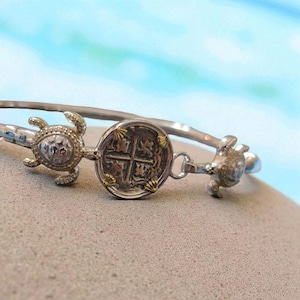 May include: Silver-toned bangle bracelet featuring a central coin design and two turtle charms. The coin has a cross pattern and is accented with gold-colored details. The turtles have textured shells and are embellished with small stones.