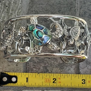 May include: A silver cuff bracelet with a detailed design of three sea turtles. The center turtle has a shell made of abalone, which is iridescent with shades of blue, green, and purple. The bracelet is 2 inches wide.