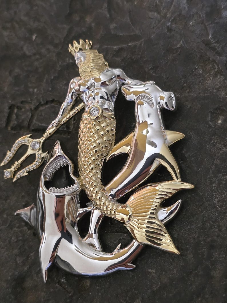 May include: A two-tone gold and silver pendant featuring a mermaid with a crown, holding a jeweled trident, and surrounded by two sharks. The mermaid's tail is gold, and the sharks are silver.