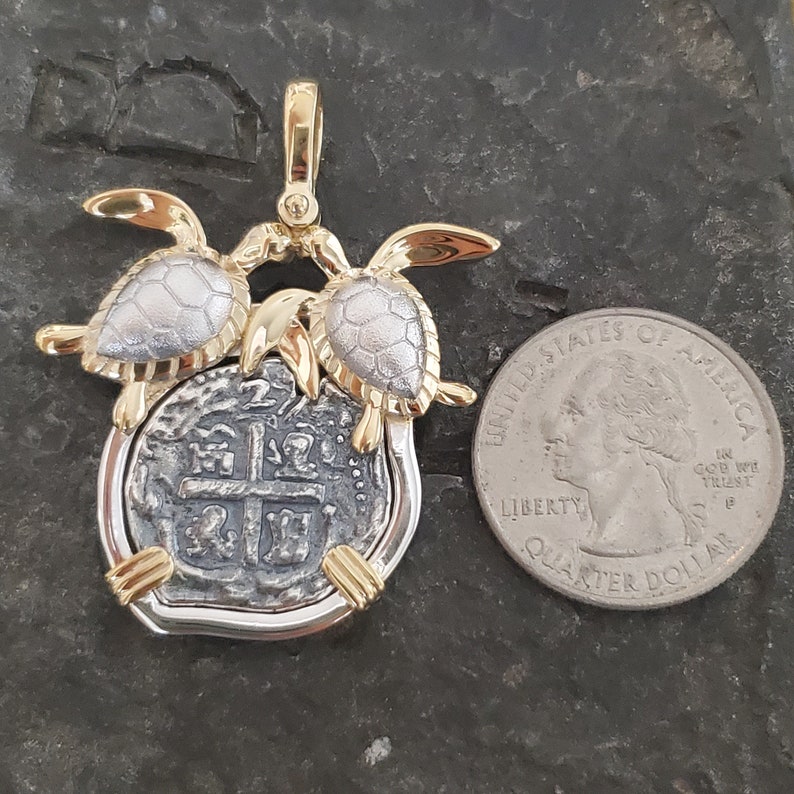 May include: A gold and silver pendant featuring a coin design with two turtle figures. The pendant has a gold bail and accents, with a silver coin center. The coin is next to a quarter for size comparison.