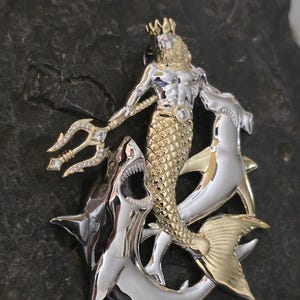 May include: A two-tone metal pendant featuring a merman with a trident, a crown, and a fish tail, flanked by two sharks. The merman is gold-colored, and the sharks are silver-colored. The pendant has a detailed design with a gemstone on the merman's chest.